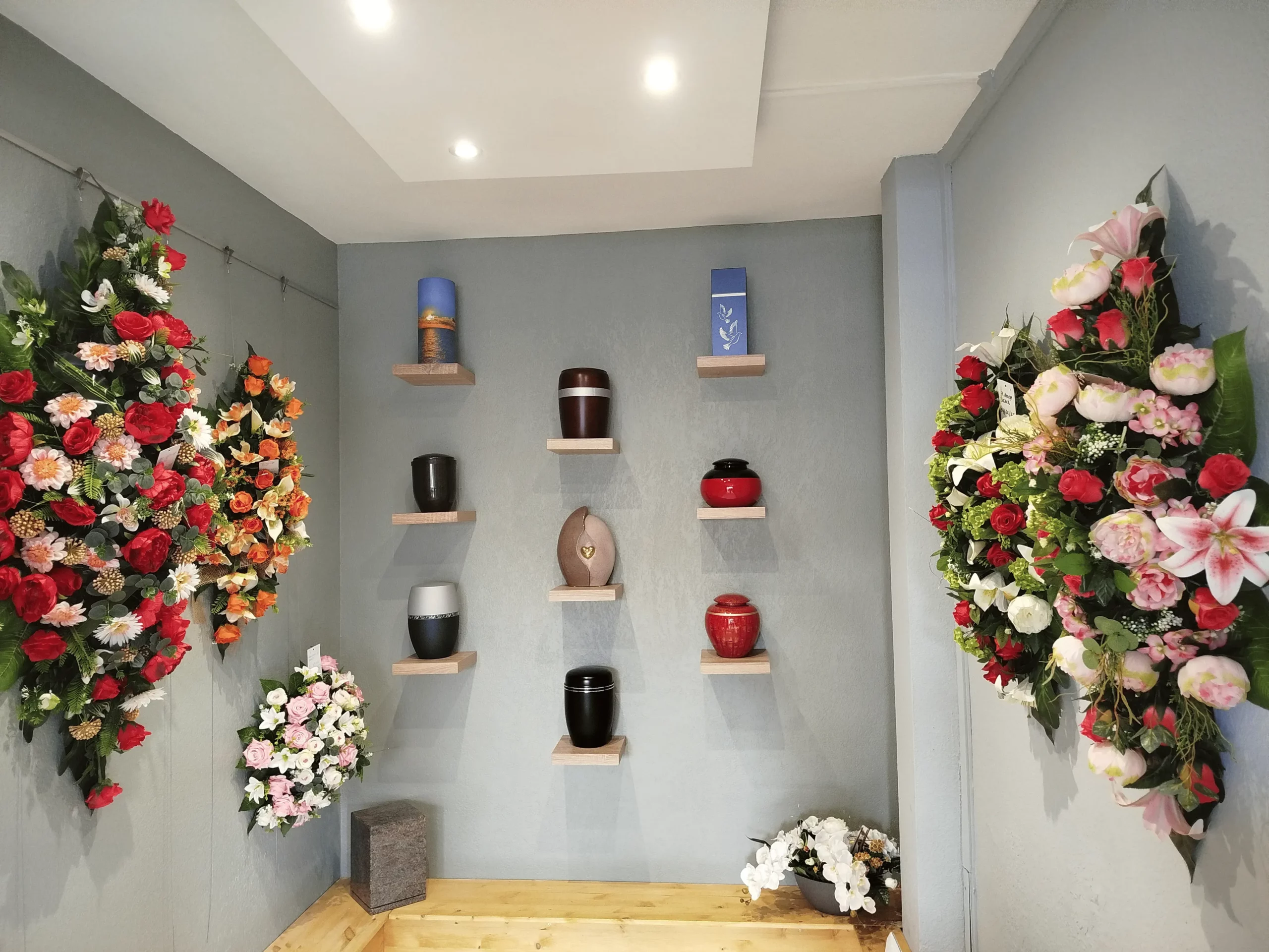 Salle d'exposition funéraire avec urnes sur étagères et gerbes de fleurs.
